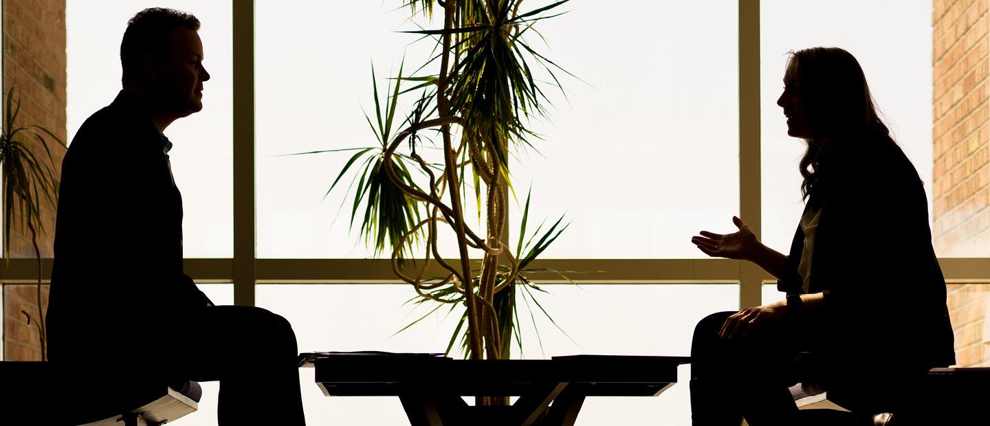The silhouette of two people seated across a table from each other, talking.