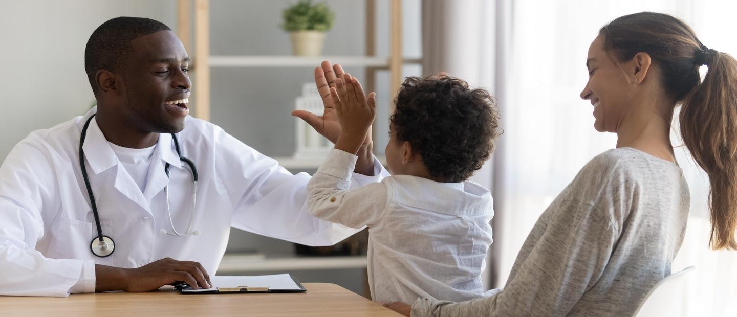 A physician's assistant giving a high five to a kid sitting in his mom's lap.