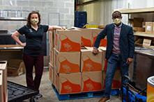 两名公开大学的员工戴着面具，站在一个仓库式的房间里，旁边放着大箱子