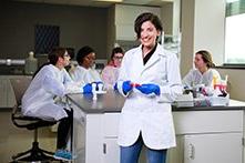阿瓦·哈迪德身穿白色实验大褂，戴着蓝色手套，在临床和诊断科学实验室里，其他学生坐在她后面.