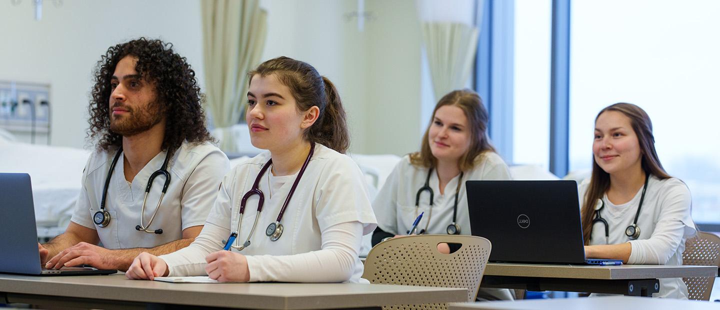 四个穿着白大褂的学生坐在一张放着听诊器的桌子前